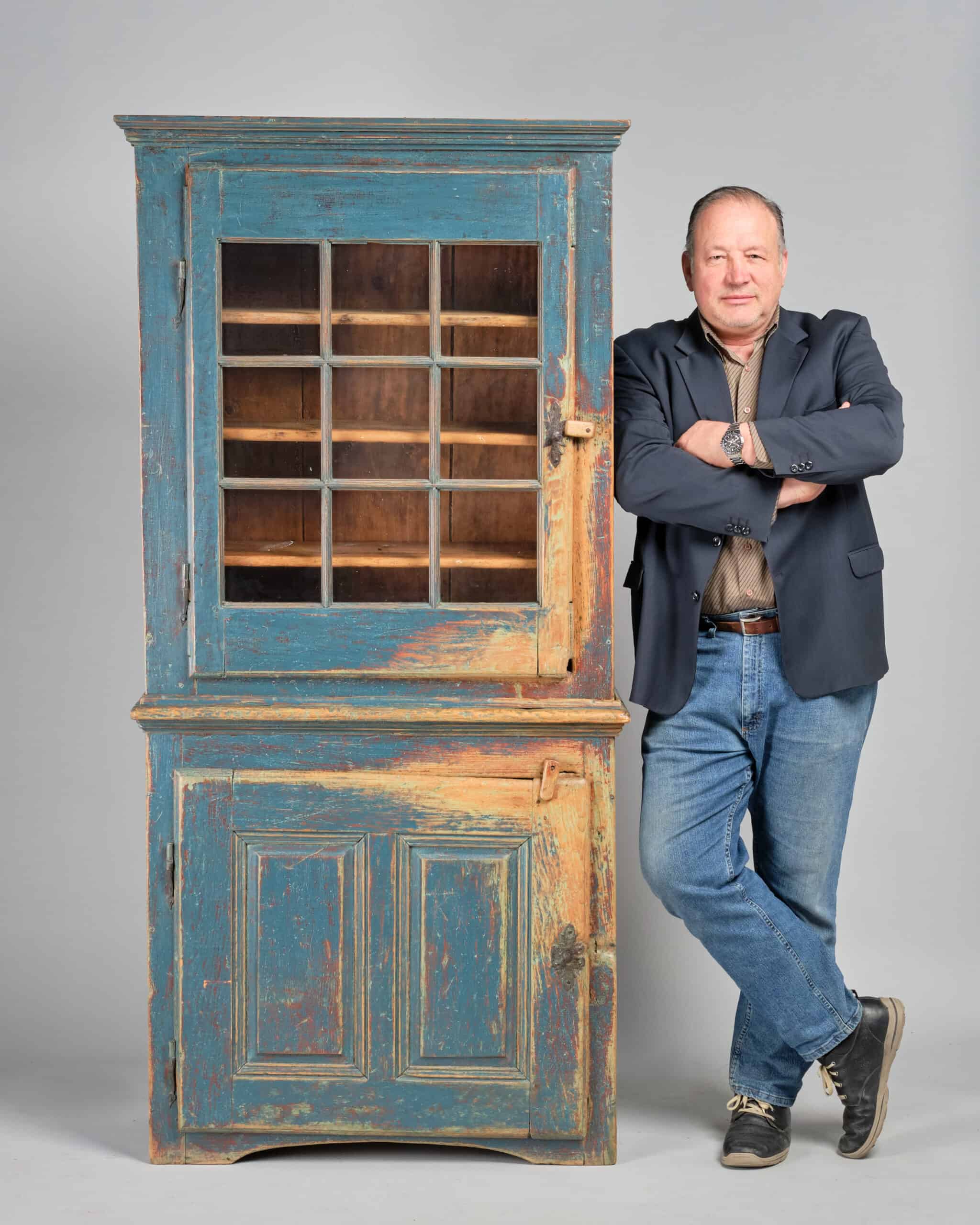 Robin Pridham, Owner of Pridham's Auctions & Appraisals leans against an old Québec cupboard that is in old blue paint over original old red paint.